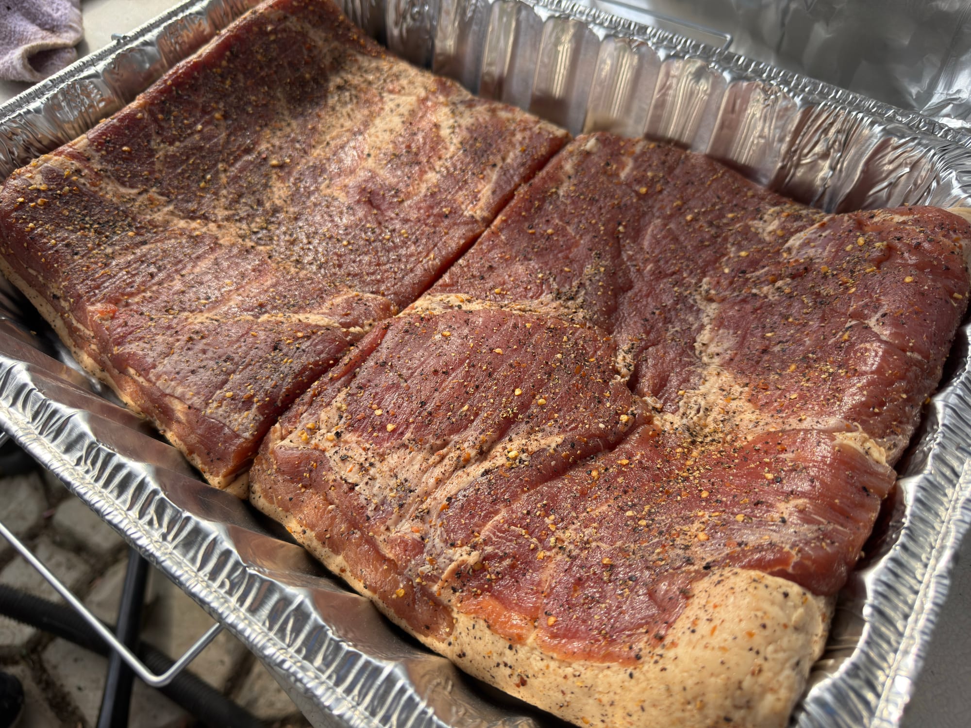 Two hunks of cured and seasoned pork belly sitting in a foil pan, ready for the smoker.