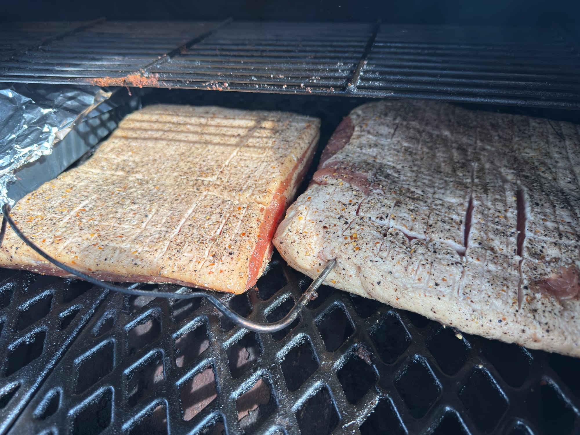 Two slabs of prepped pork belly with temperature probes in them on a pellet grill.