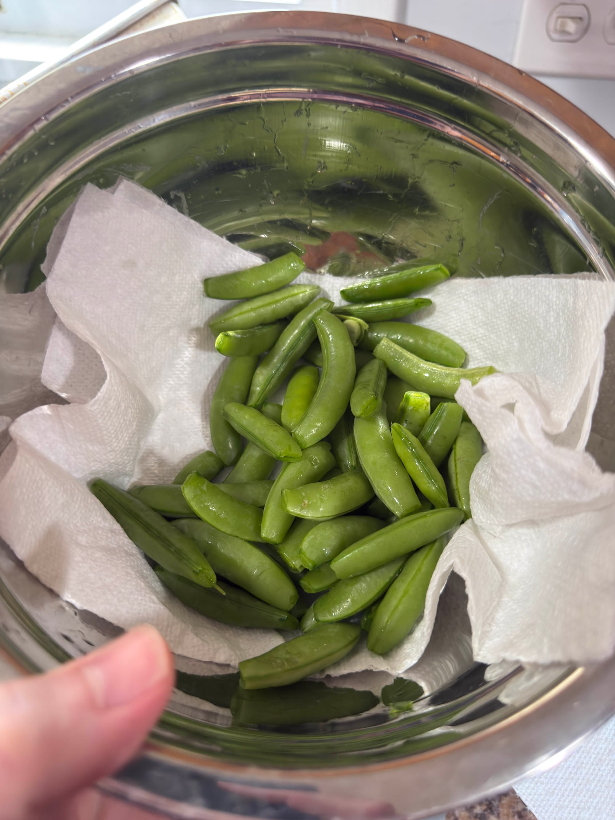 A stainless steel bowl fill of freshly-trimmed and rinsed snap peas drying on a paper towel.