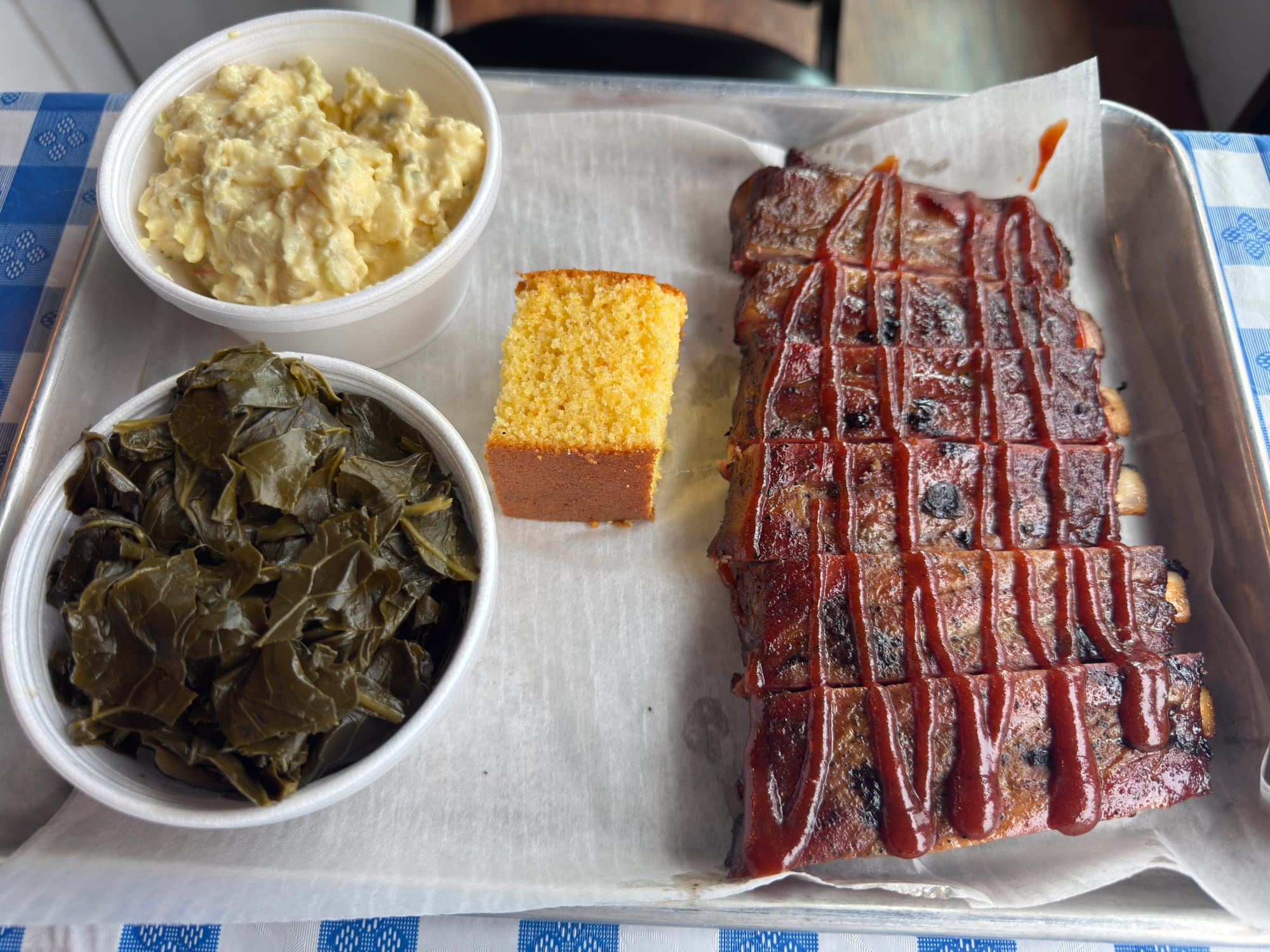 A serving tray full of smoked ribs, corn bread, potato salad, and greens.