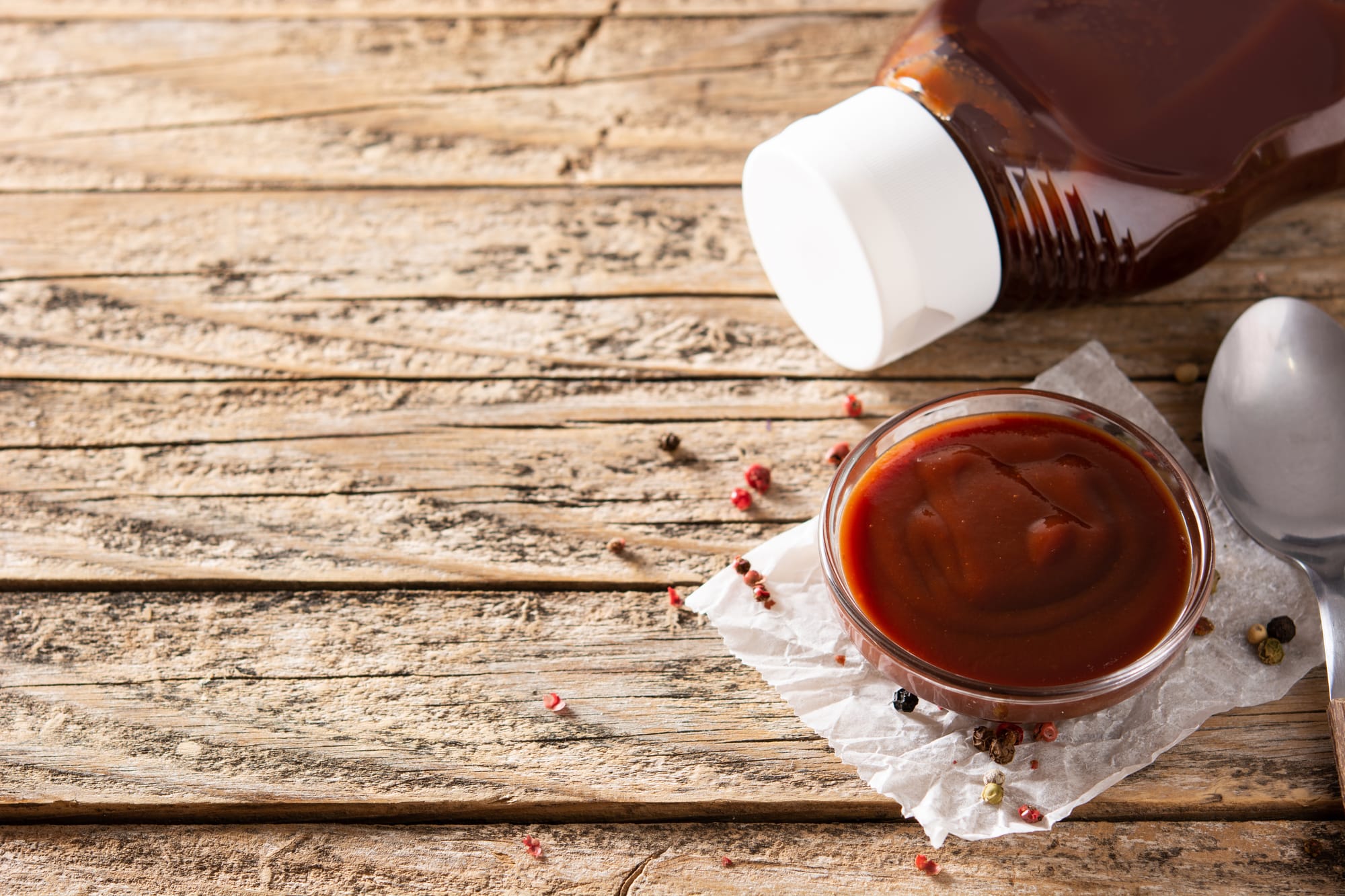 Barbecue sauce in a glass bowl on a table with the bottle of sauce and a spoon nearby.