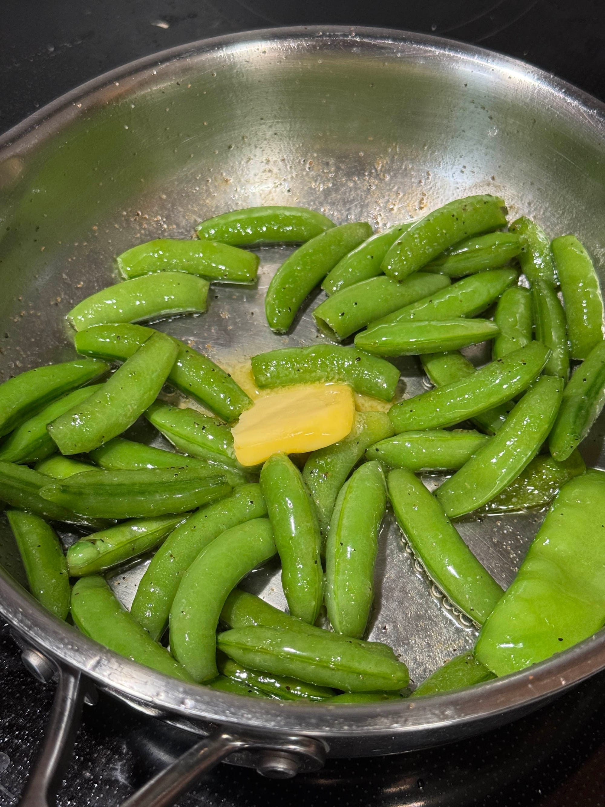 A stainless steel skillet full of fried snap peas with a dollop of Kerrygold butter melting on top.